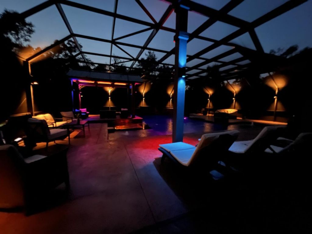 An evening photo of ambience lighting mounted on the support posts of a screened in pool enclosure around the pool.