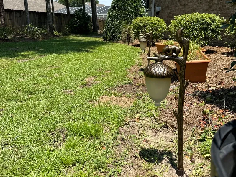 A photo of a landscape path light shaped like a tree branch and acorn with a squirrel sitting on top on the edge of a flower bed.